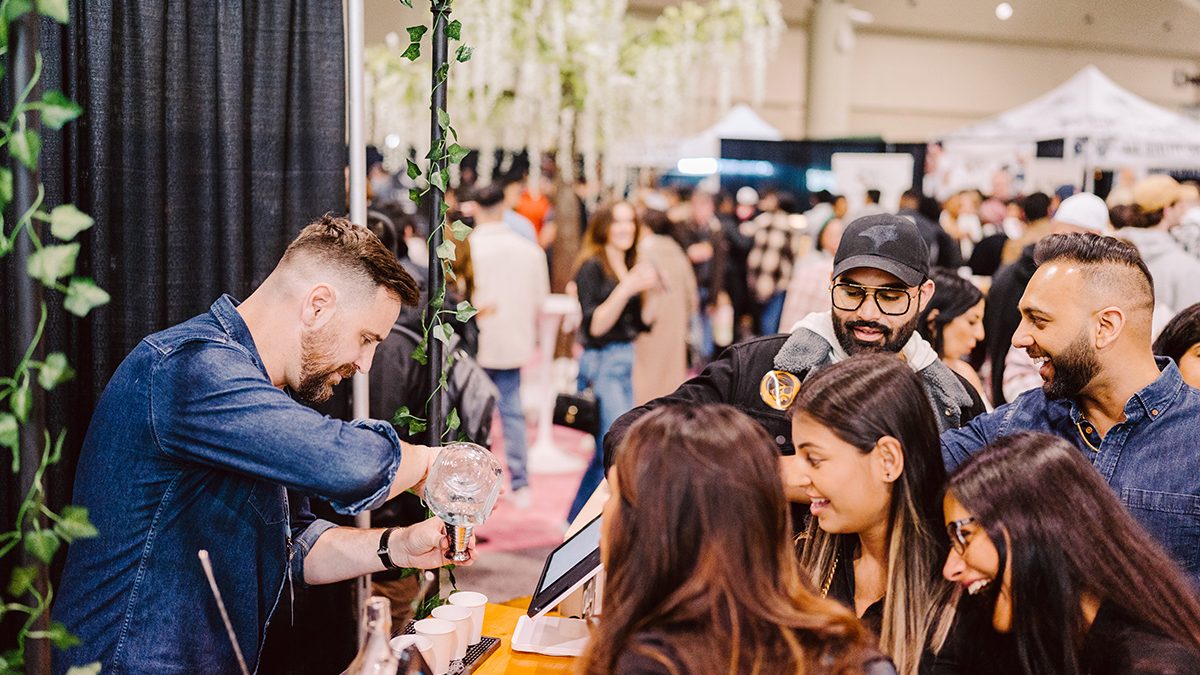 A vendor serves drinks from a counter amidst a crowded event space decorated with greenery.