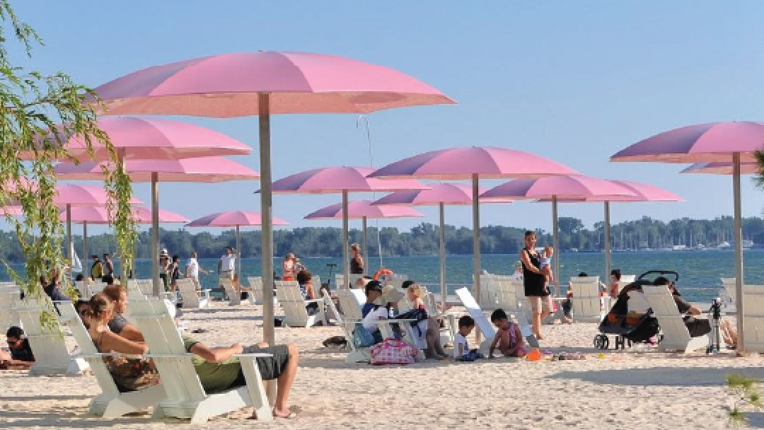 Beach scene with pink umbrellas providing shade, while people relax on lounge chairs by the water on a sunny day.