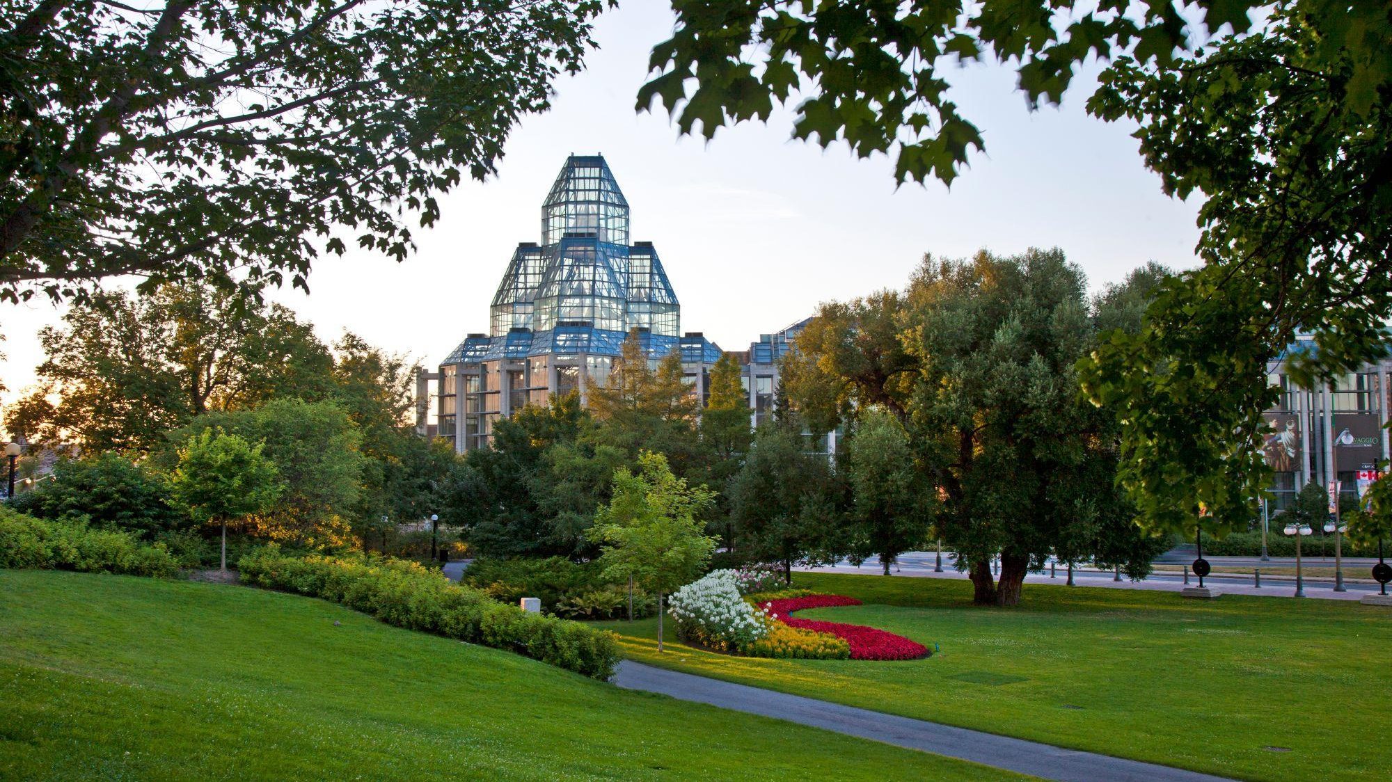 A lush park with colorful flower beds in the foreground, featuring a modern glass pyramid building in the background during sunset.