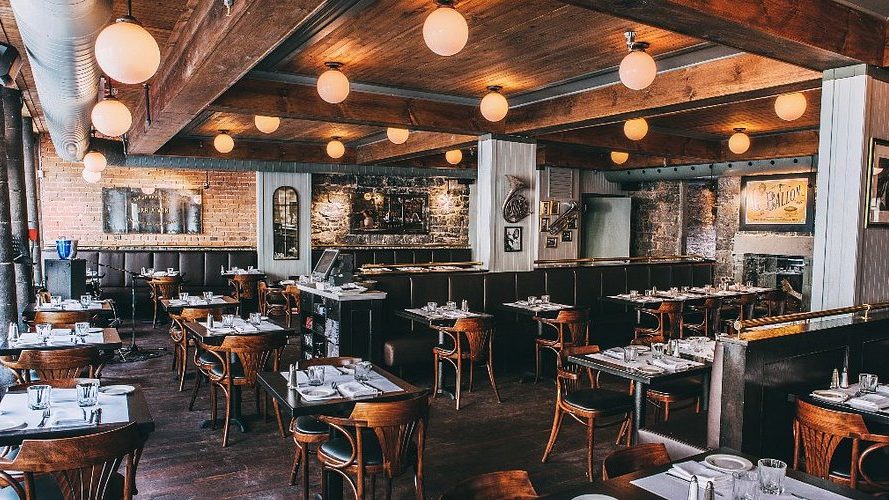 Cozy restaurant interior with wooden tables and chairs, warm lighting from round ceiling lights, exposed brick walls, and elegant table settings.