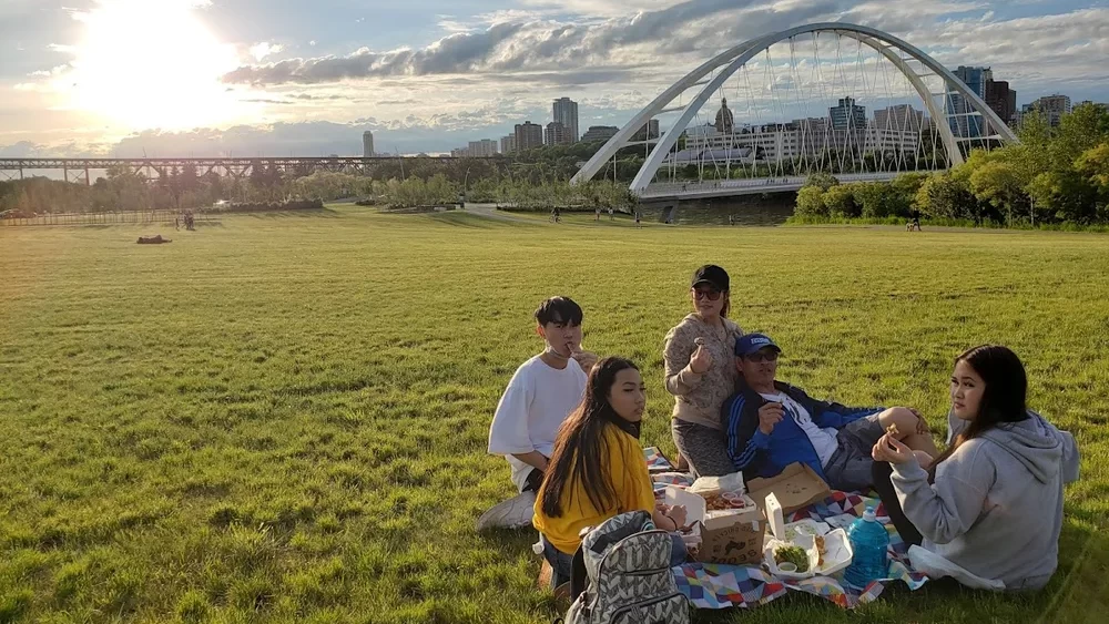 A group enjoys a picnic on a sunny day in a green park, with a bridge and Edmonton skyline in the background.