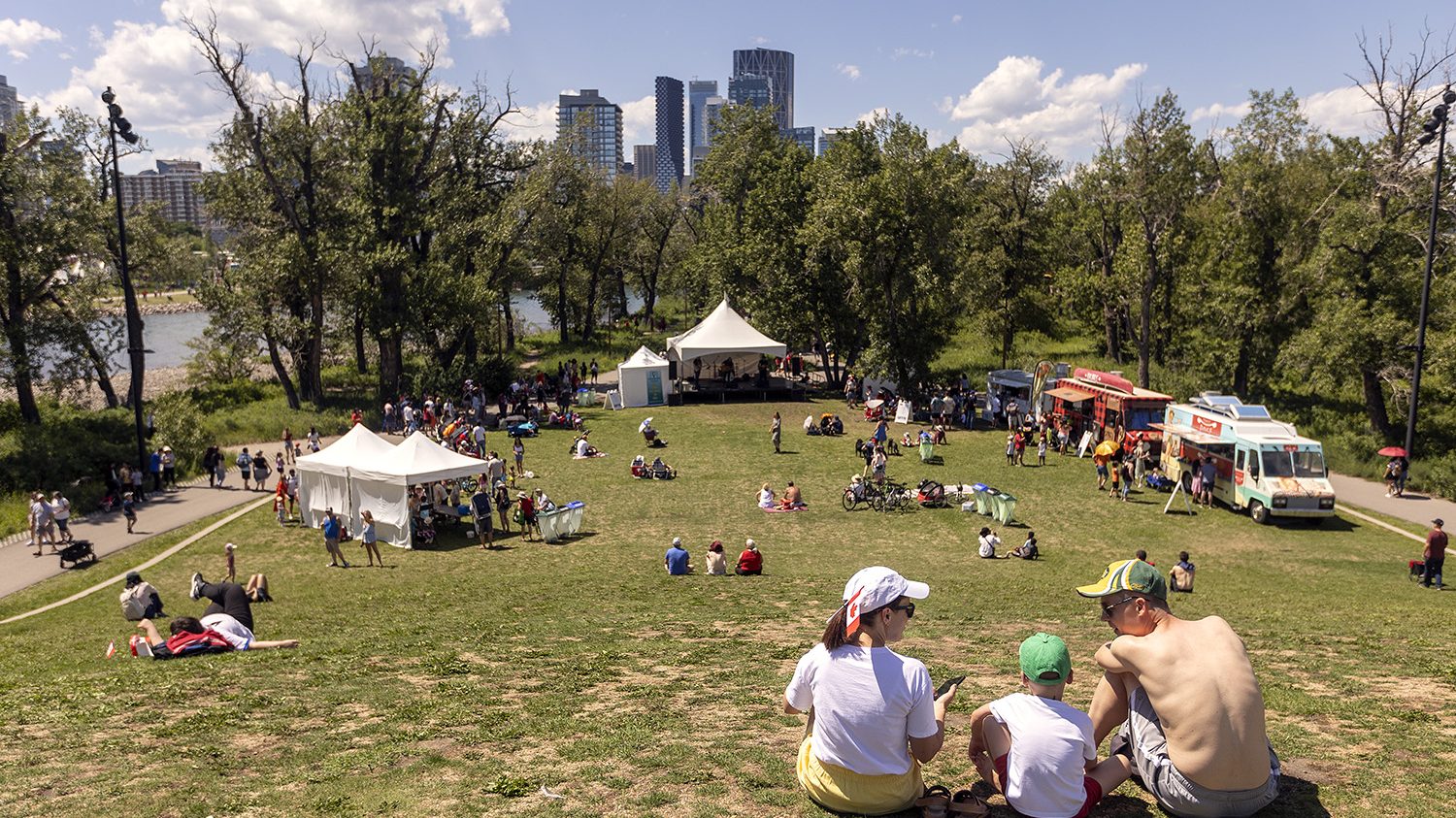 A sunny park scene filled with families and friends enjoying food trucks, entertainment tents, and recreational activities by the water.