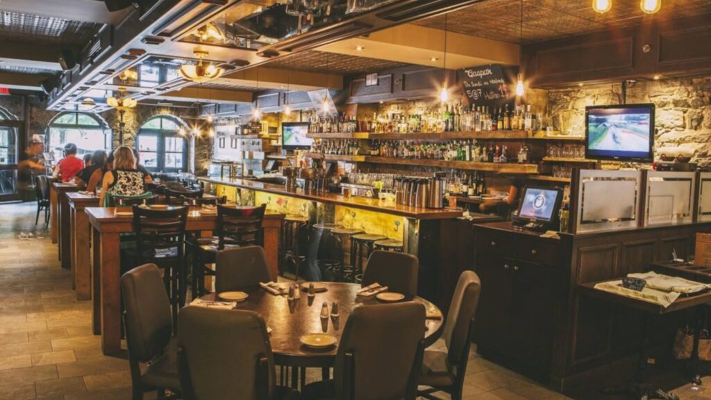 Cozy pub interior with rustic stone walls, wooden tables, and a well-stocked bar. Warm lighting creates an inviting ambiance; patrons relax at tables.