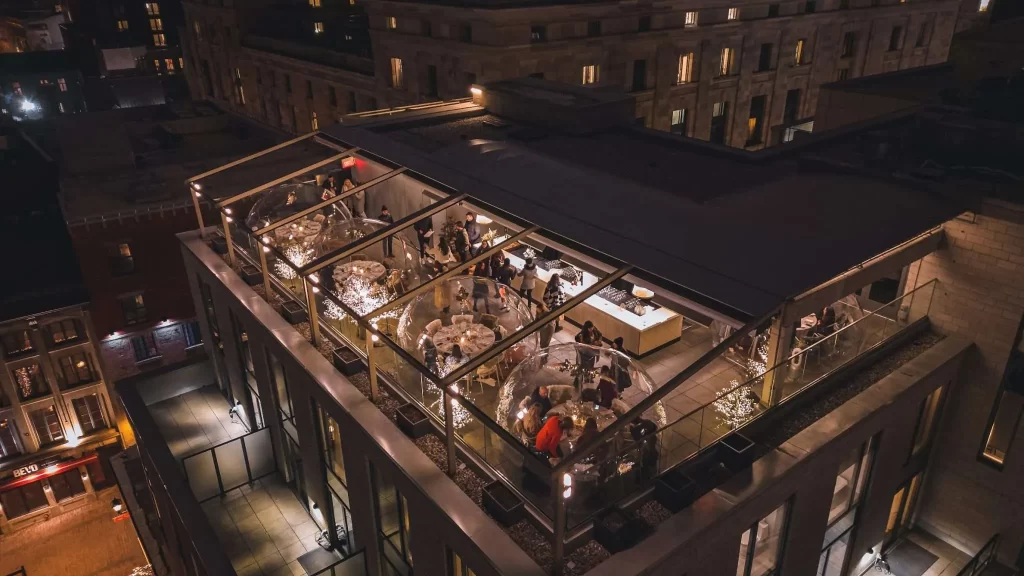 Aerial view of a rooftop restaurant at night, with groups dining in transparent igloos. Warm lighting creates an inviting ambiance amidst an urban cityscape.