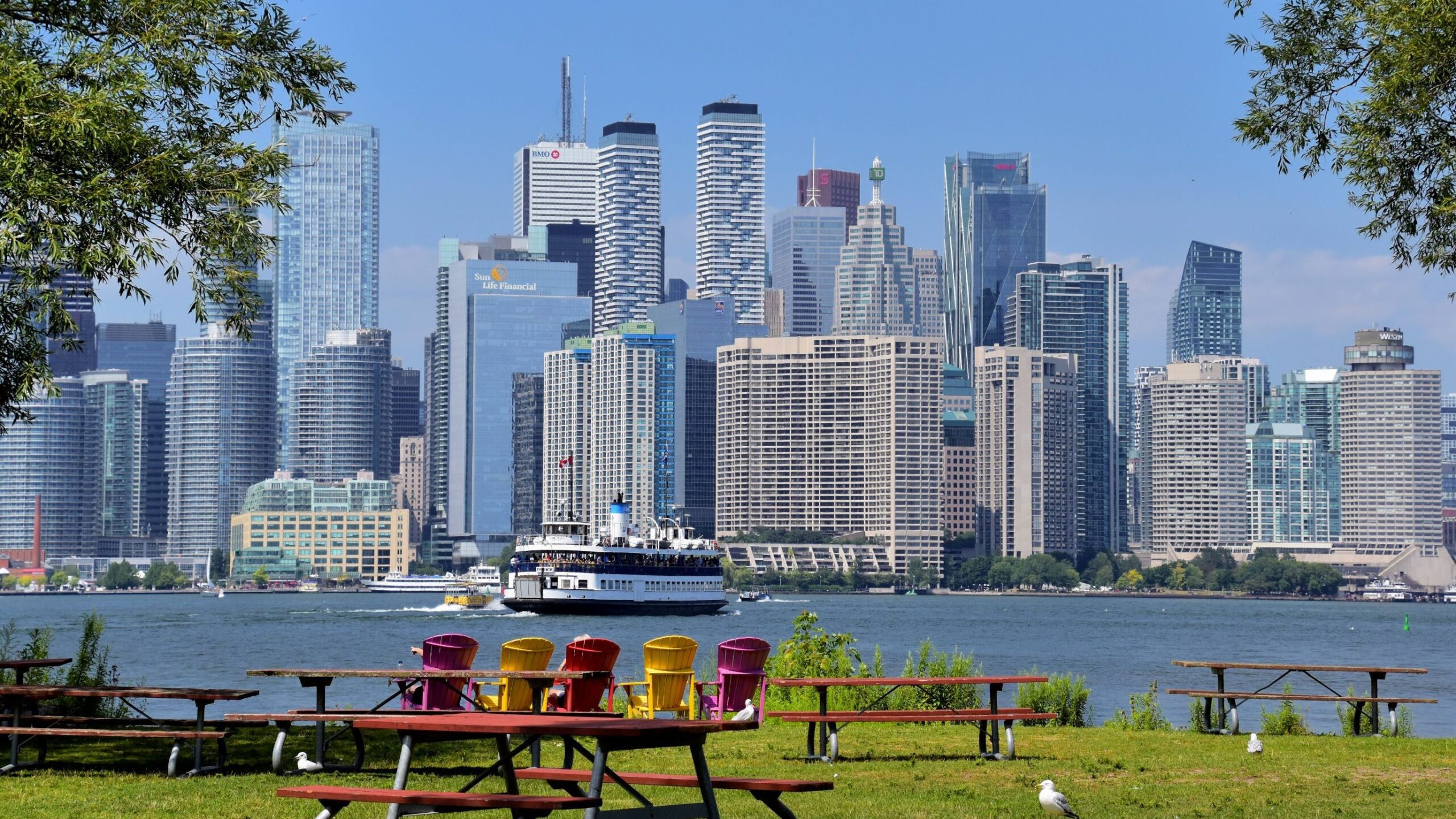 Colorful chairs and picnic tables in a park with a view of a bustling city skyline along the waterfront on a sunny day.