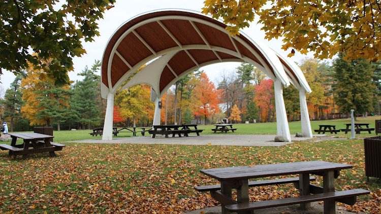 An open pavilion with a curved roof surrounded by picnic tables and colorful autumn foliage in a park setting.