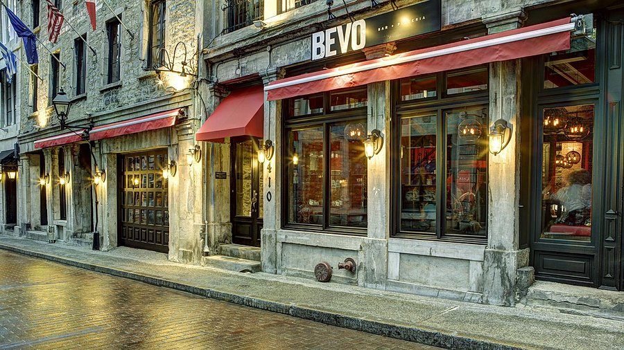 Charming cobblestone street scene with warm lighting. "BEVO" bar features large windows, red canopies, and rustic stone facade, creating an inviting ambiance.