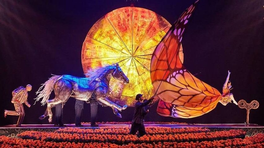 A dynamic scene from LUZIA featuring a puppet horse in motion, surrounded by performers in vibrant, colorful costumes. A glowing orange wheel and butterfly wings enhance the magical atmosphere, set against a backdrop of vivid flowers.