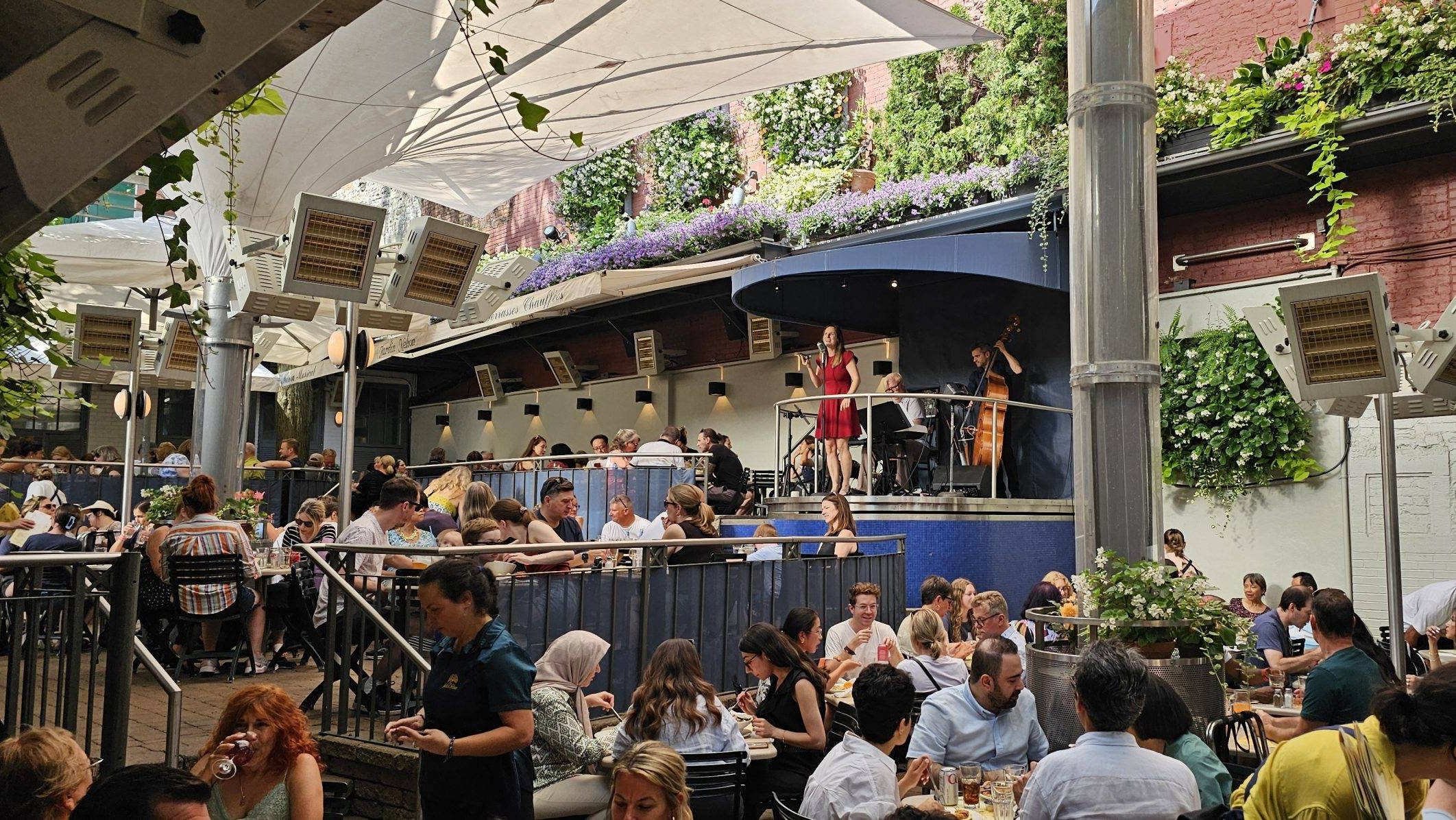 Bustling outdoor restaurant scene with a live band performing on stage. Diners enjoy meals under large white canopies amid vibrant greenery. Lively and relaxed atmosphere.