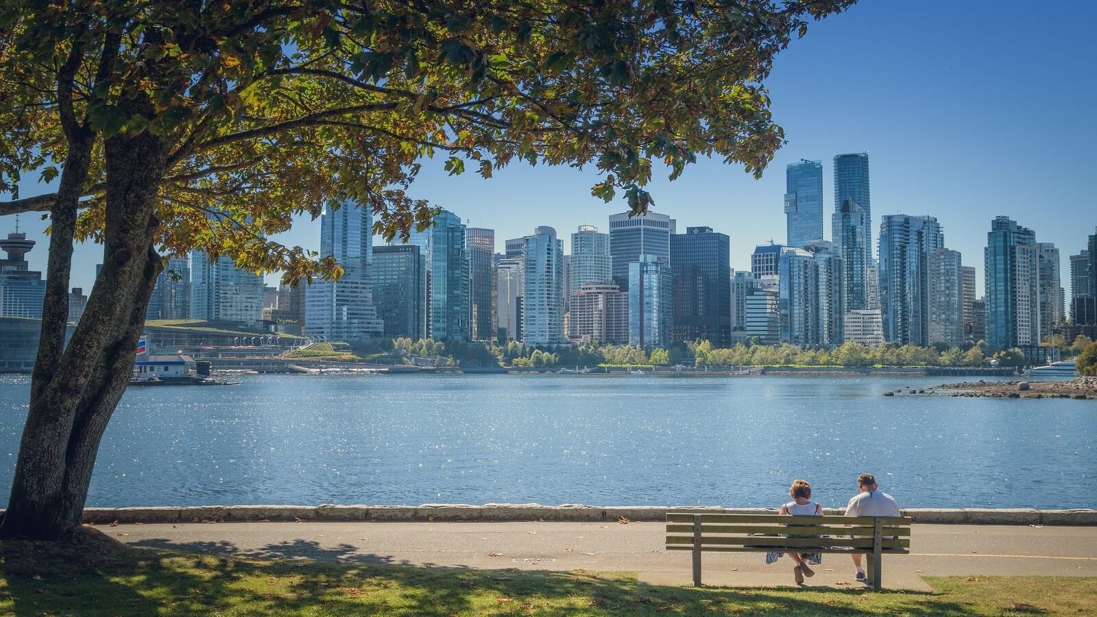 A couple sits on a bench by the water, enjoying a sunny day with Vancouver's skyline in the background and trees framing the scene.