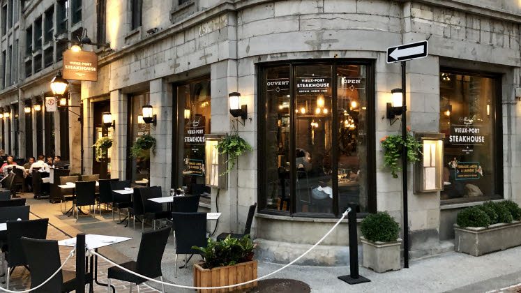 Street view of Vieux-Port Steakhouse with outdoor seating. Elegant stone façade, warm lighting from inside, and hanging plants create a welcoming ambiance.