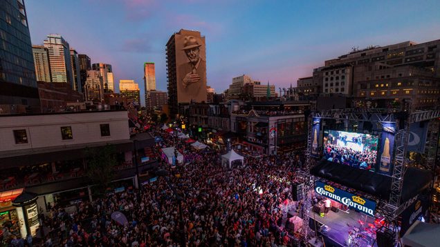 Une foule dynamique se rassemble lors d'un concert en plein air sous un ciel coloré, avec un grand écran et la ligne d'horizon de Montréal en arrière-plan.