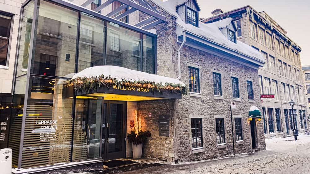 L'entrée de l'hôtel William Gray présente une façade en verre moderne adossée à un bâtiment historique en pierre, orné d'un décor hivernal et de neige.