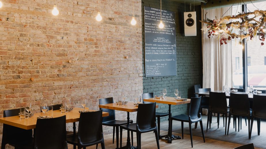 Un restaurant moderne avec des tables en bois, des chaises noires, des murs en briques apparentes, un menu au tableau noir et des suspensions lumineuses.