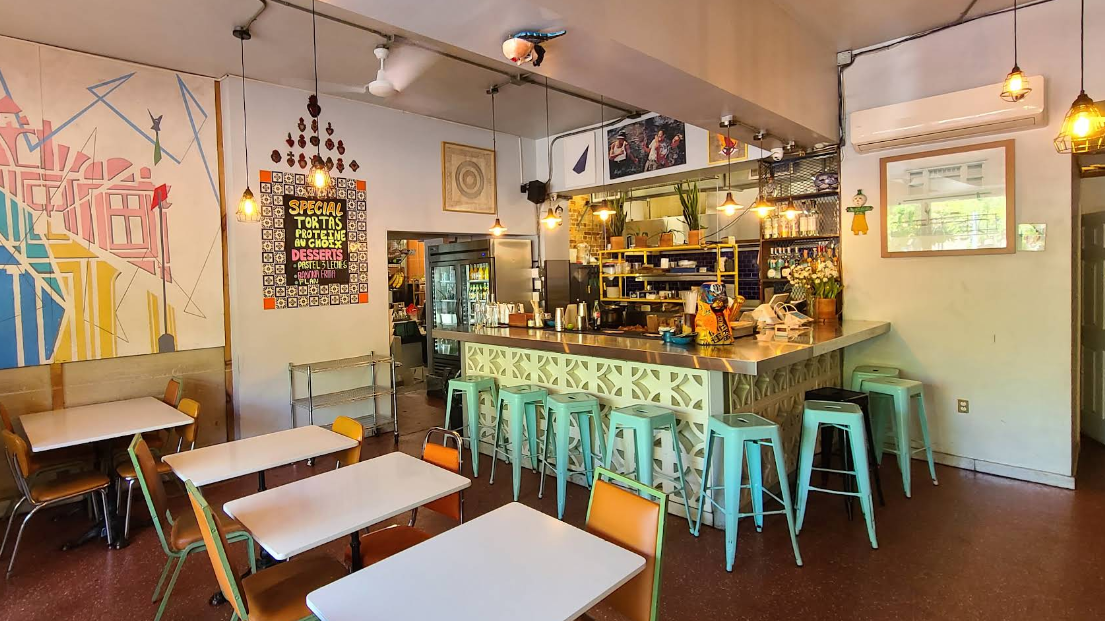 Un intérieur de taqueria chaleureux avec une fresque murale vibrante, un bar avec des tabourets bleu sarcelle, des tables et un éclairage chaleureux.