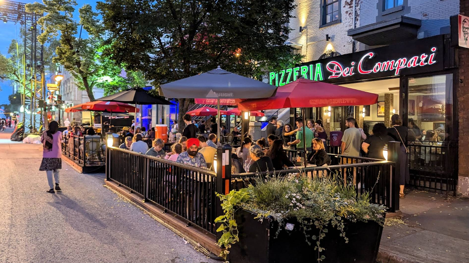 Une scène de restauration en plein air animée à la Pizzeria Dei Compari, avec des clients savourant leurs repas sous des parasols colorés le soir.