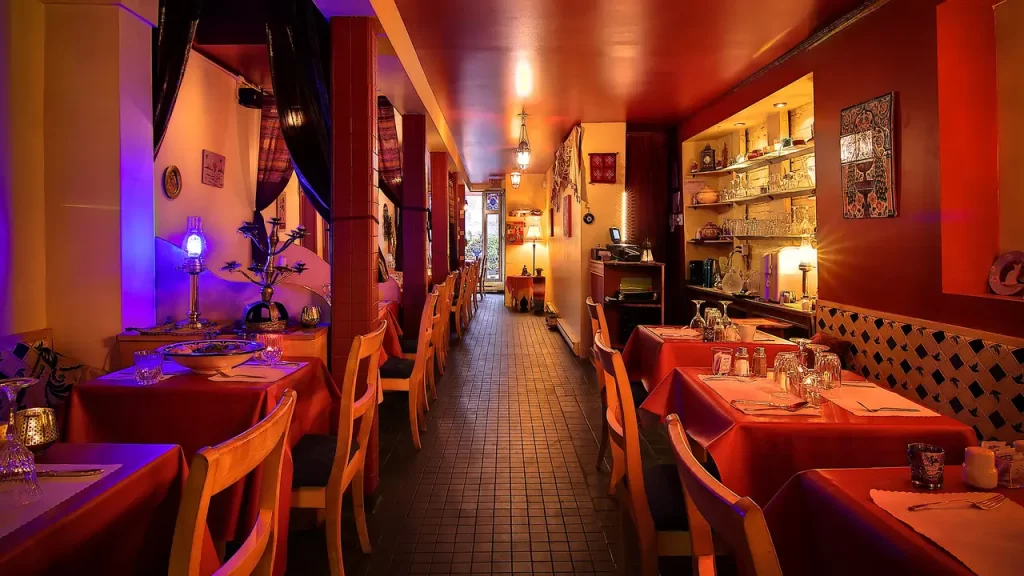 Intérieur de restaurant chaleureux avec nappes rouges, chaises en bois et éclairage d'ambiance, menant à une entrée chaleureusement éclairée.