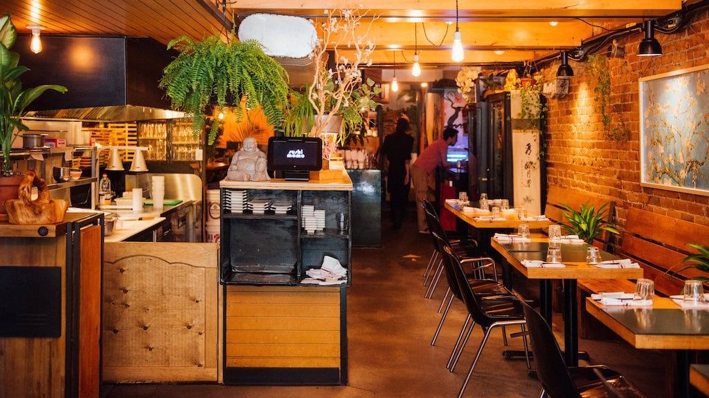 Intérieur de restaurant chaleureux avec décor en bois, plantes et tables dressées pour les repas, avec un coin cuisine en arrière-plan et un éclairage chaleureux.