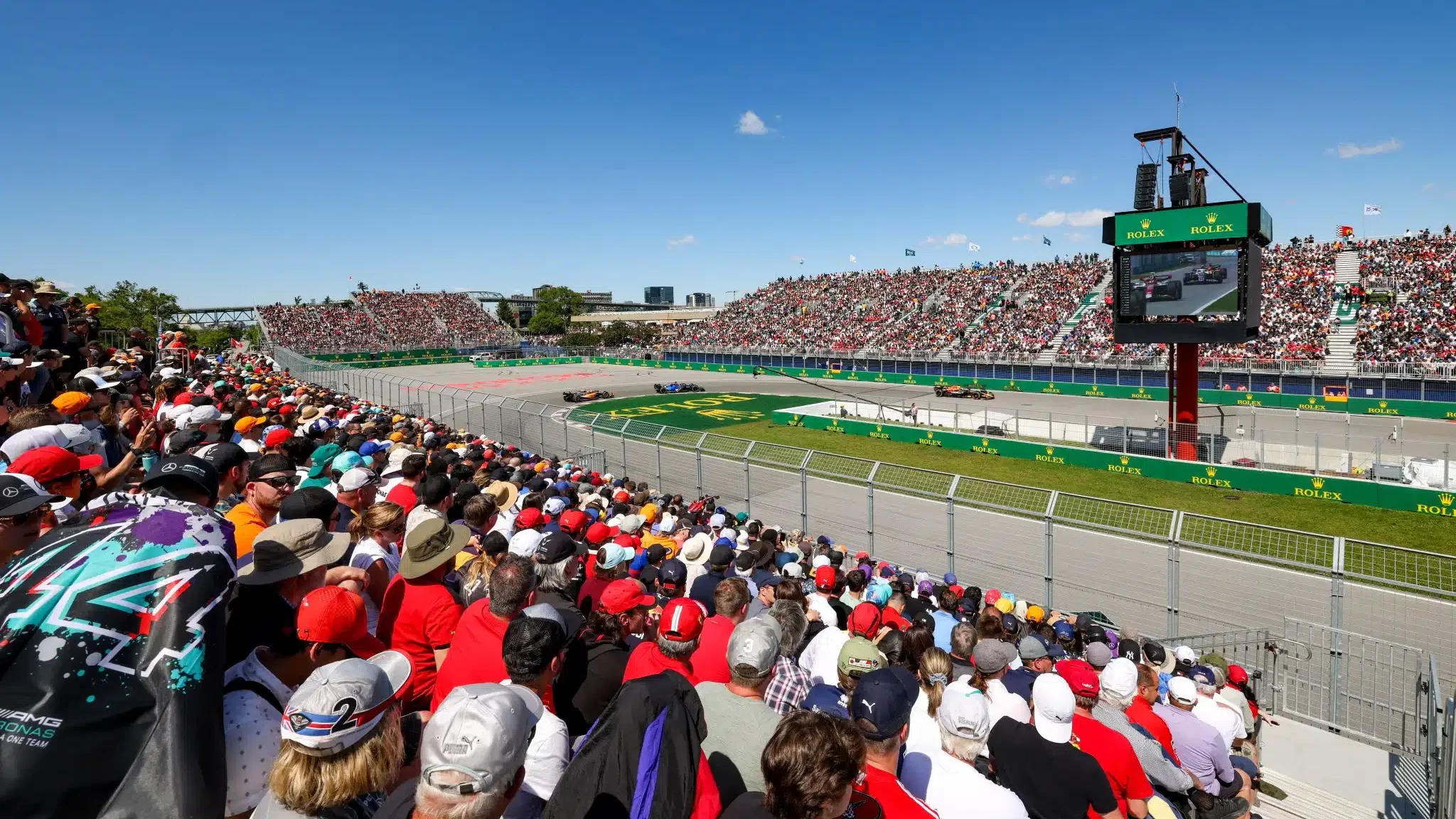 Des tribunes remplies de spectateurs surplombent le circuit de Formule 1 du Grand Prix du Canada, où les voitures de course filent sous un ciel bleu clair et où un grand tableau d'affichage se profile en arrière-plan.